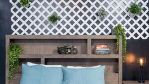 A bed with blue pillows, a wooden nightstand with a lamp, a lattice wall with plants, and a shelf with books and a toy car. - Charming apt -2 Bedroom -Close to Beach/Sleeps 6 (Ipswich)