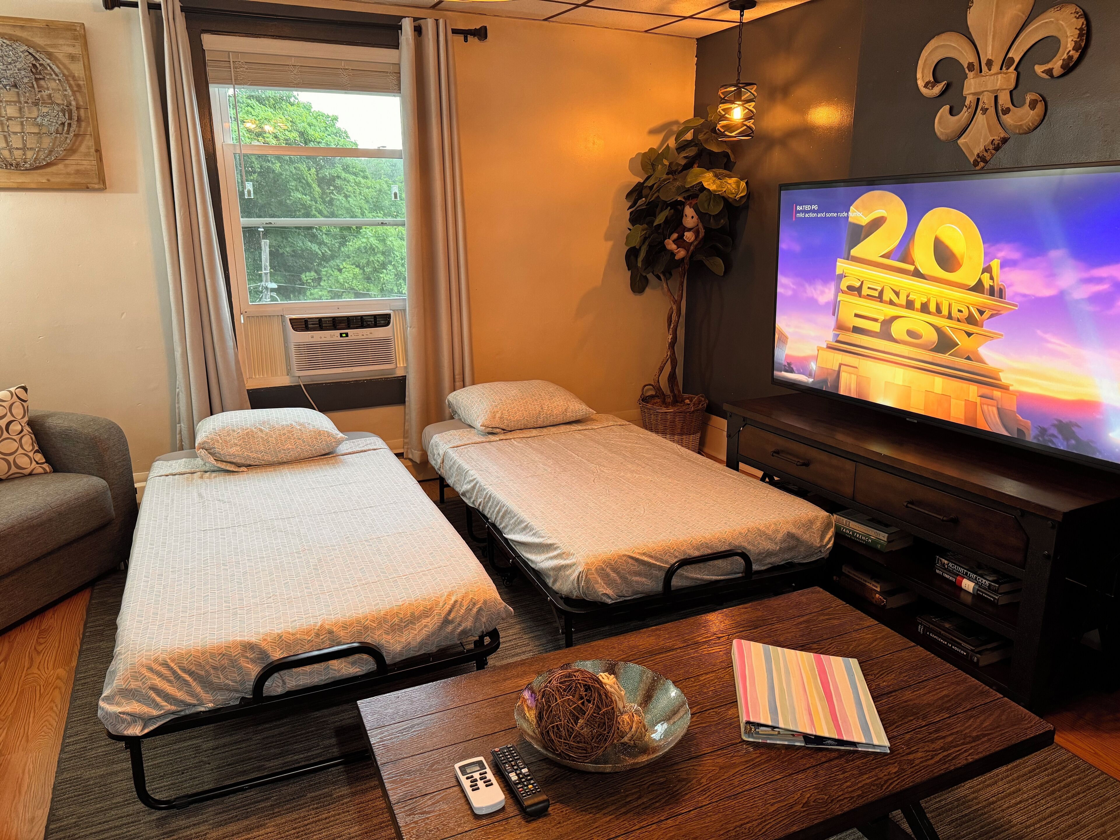 Une chambre d’hôtel avec deux lits simples, un téléviseur affichant le logo « 20TH CENTURY FOX », une table basse en bois avec une télécommande, un livre et une plante décorative.