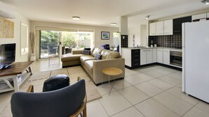 Una sala moderna con un sofá, televisor y comedor. La cocina tiene armarios blancos y un refrigerador.