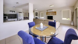 Una sala de estar moderna con una mesa redonda de vidrio, sillas azules, un sofá y una cocina con refrigerador y lavamanos.