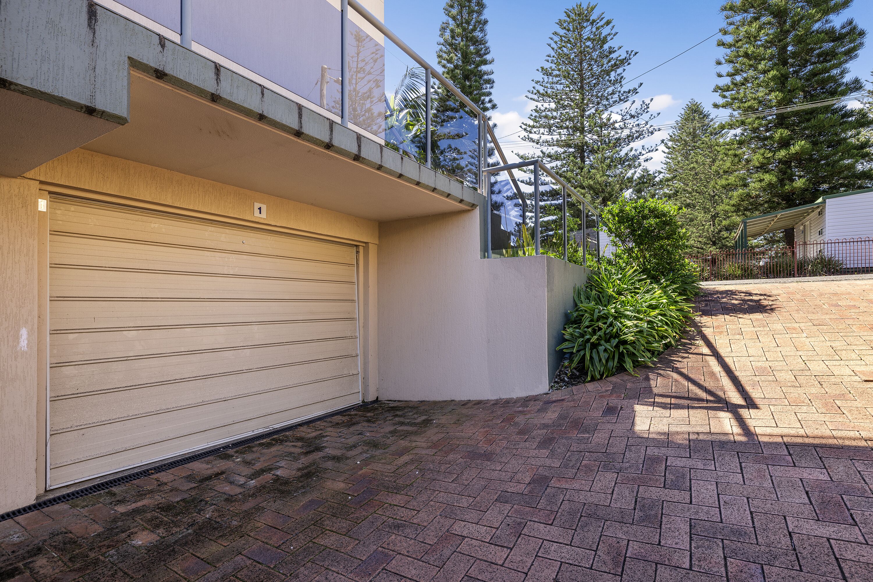 A residential garage with a closed garage door, a brick pathway leading to the entrance, and a small garden area with greenery.