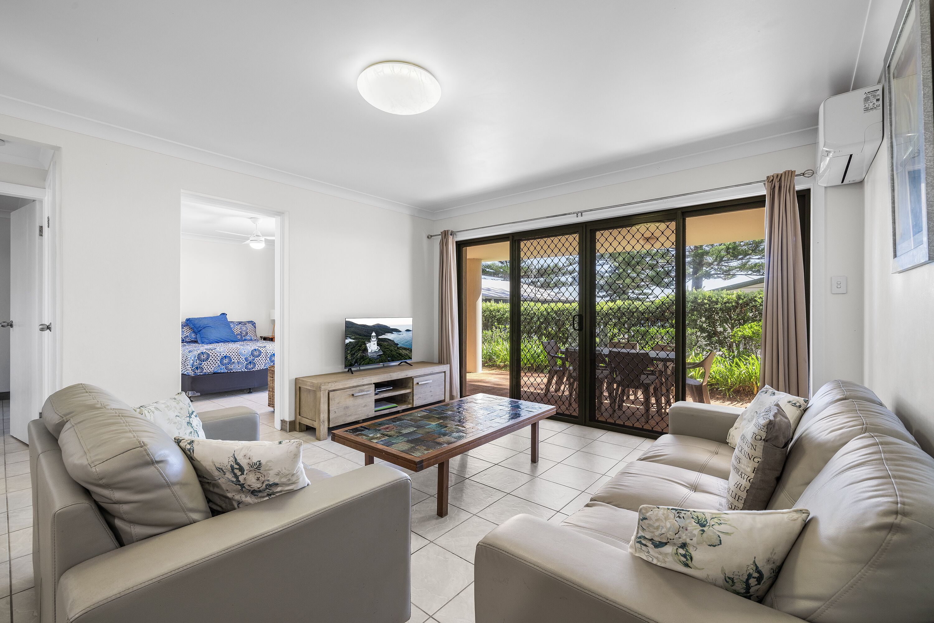 A modern living room with a sofa, a coffee table, and a TV. There is a sliding glass door leading to a balcony with a view of greenery.