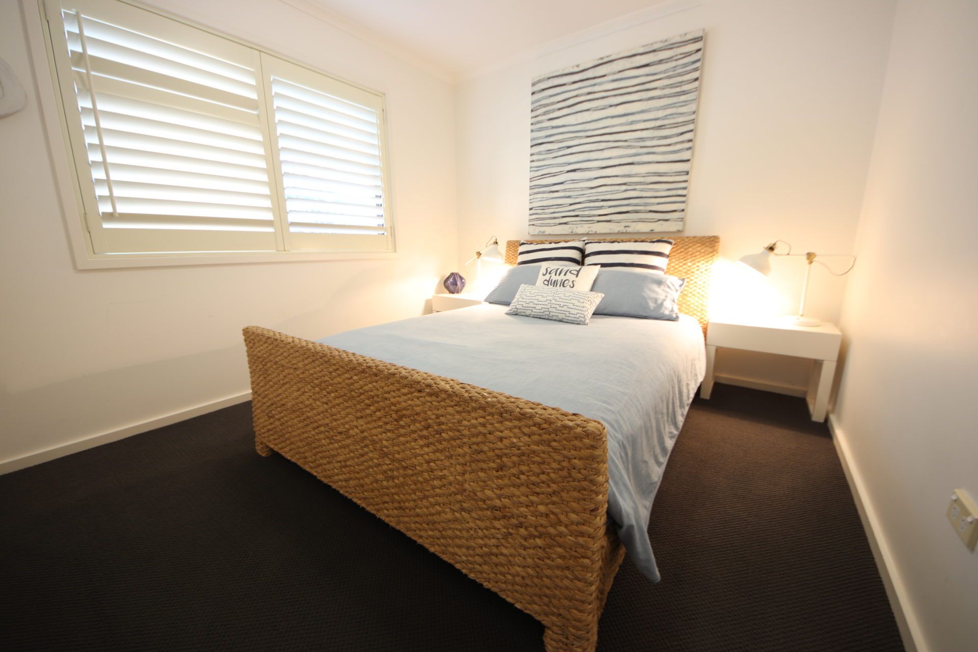 A bedroom with a large bed, two bedside tables, a wall-mounted artwork, and a window with blinds.
