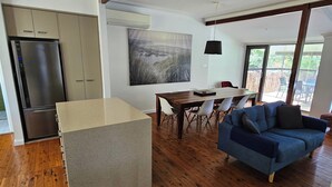 A modern kitchen with a built-in refrigerator, a dining area with a wooden table and chairs, and a blue sofa in a living area with large windows. - Comfortable air conditioned, pet friendly holiday home with large open... (South West Rocks)