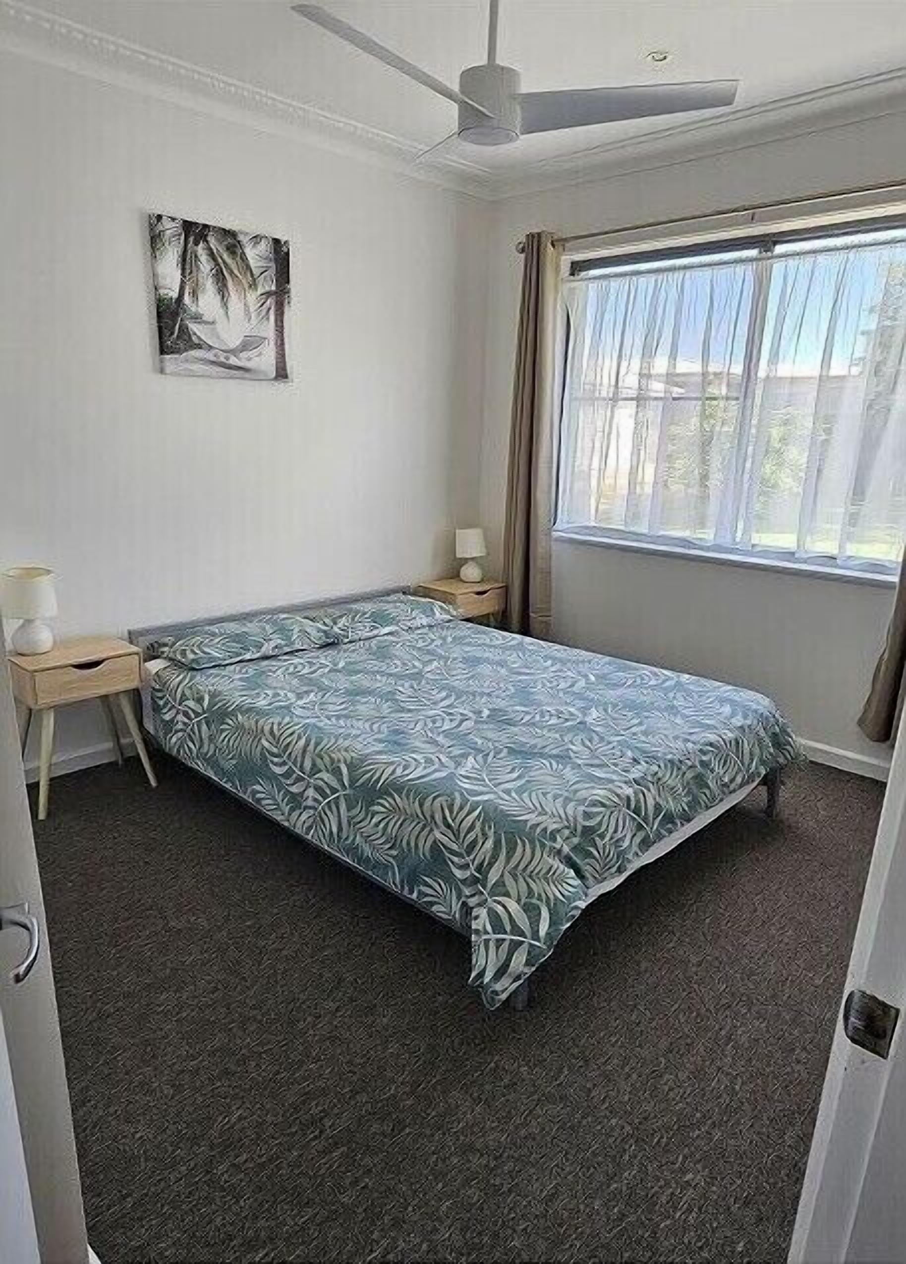 Un lit simple dans une chambre dotée d’un ventilateur de plafond, d’une table de chevet avec une lampe, d’une fenêtre avec des rideaux et d’un tableau accroché au mur.