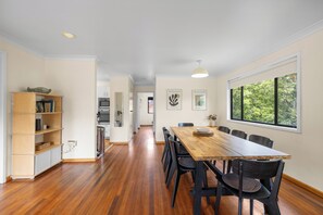 Un comedor con mesa y sillas de madera, una estantería y una ventana con vistas a árboles.