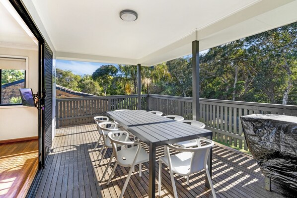 A covered outdoor deck with a wooden table and white chairs, surrounded by a wooden railing and a view of trees.