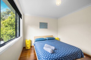 A bedroom with a large window, a bed with blue patterned bedding, a wooden headboard, and a yellow bedside table with a lamp.