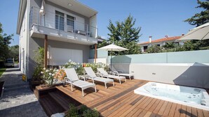 Ett modernt hus i två våningar med en träveranda, utomhusmöbler och en bubbelpool.