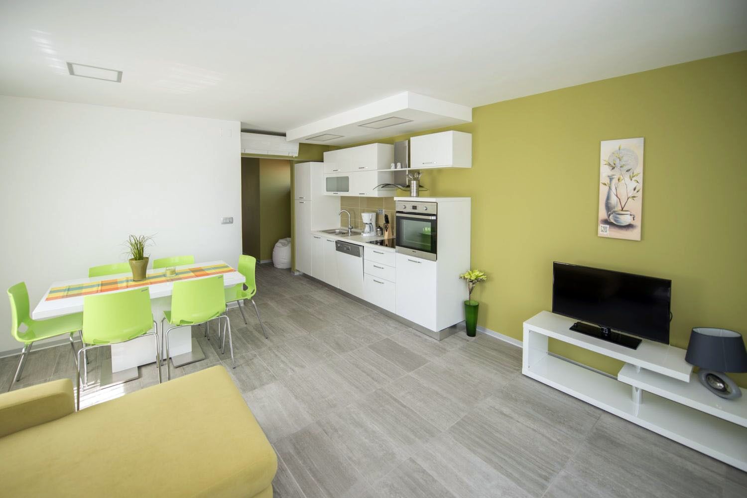 A modern kitchen with white cabinets, a dining area with green chairs, and a TV on a white stand.