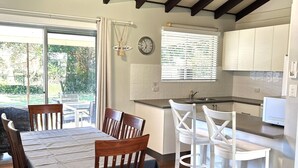 A dining area with wooden chairs and a table, a kitchen with white cabinets, and a clock on the wall.