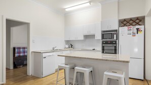 A modern kitchen with white cabinets, a microwave, an oven, and a refrigerator. There are two white stools and a small island with a sink. - Beach Break - 22 Bay St Hat Head (Hat Head)