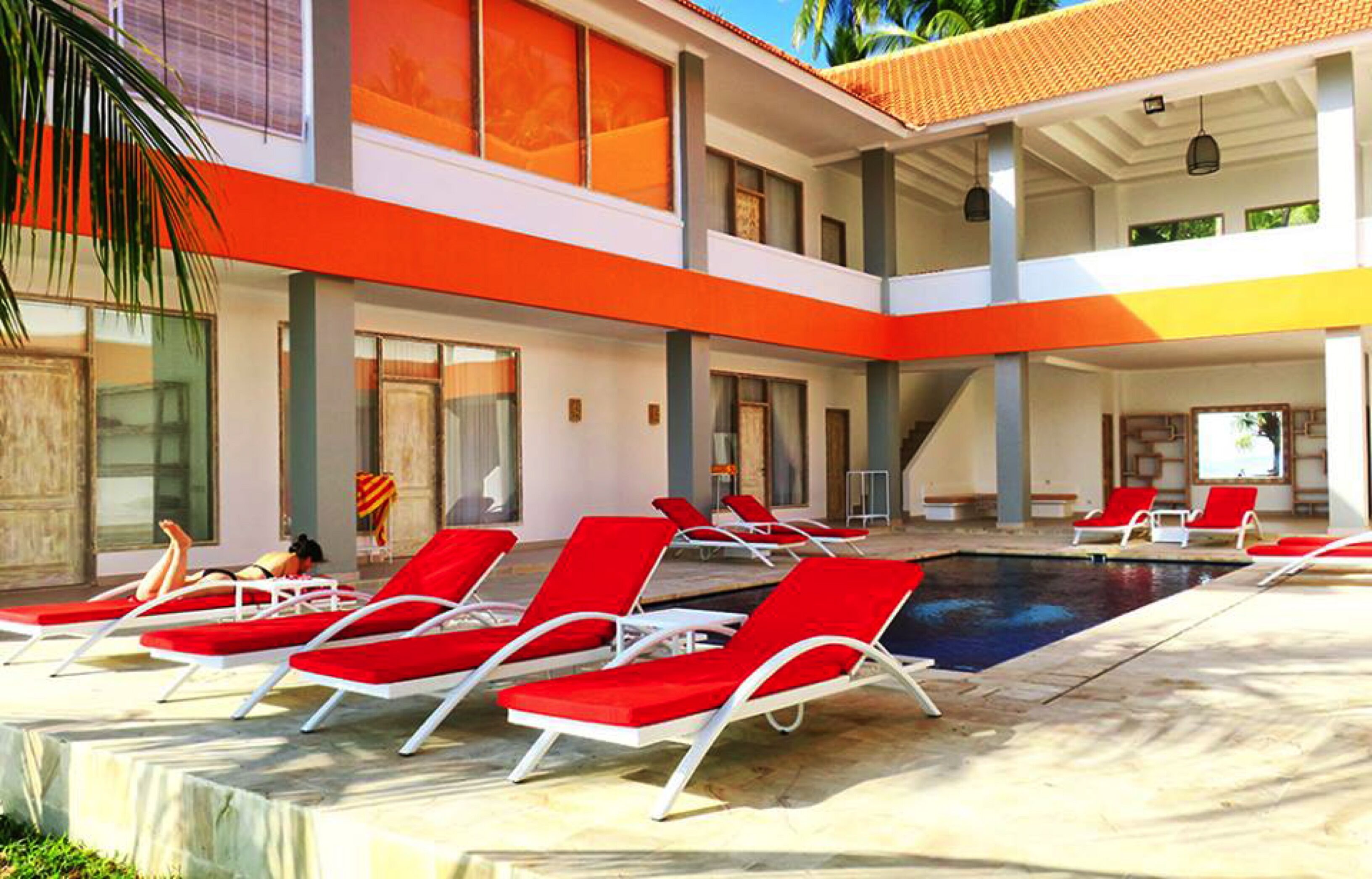 A pool area with red lounge chairs, a clear pool, and a modern building with orange and white colors.