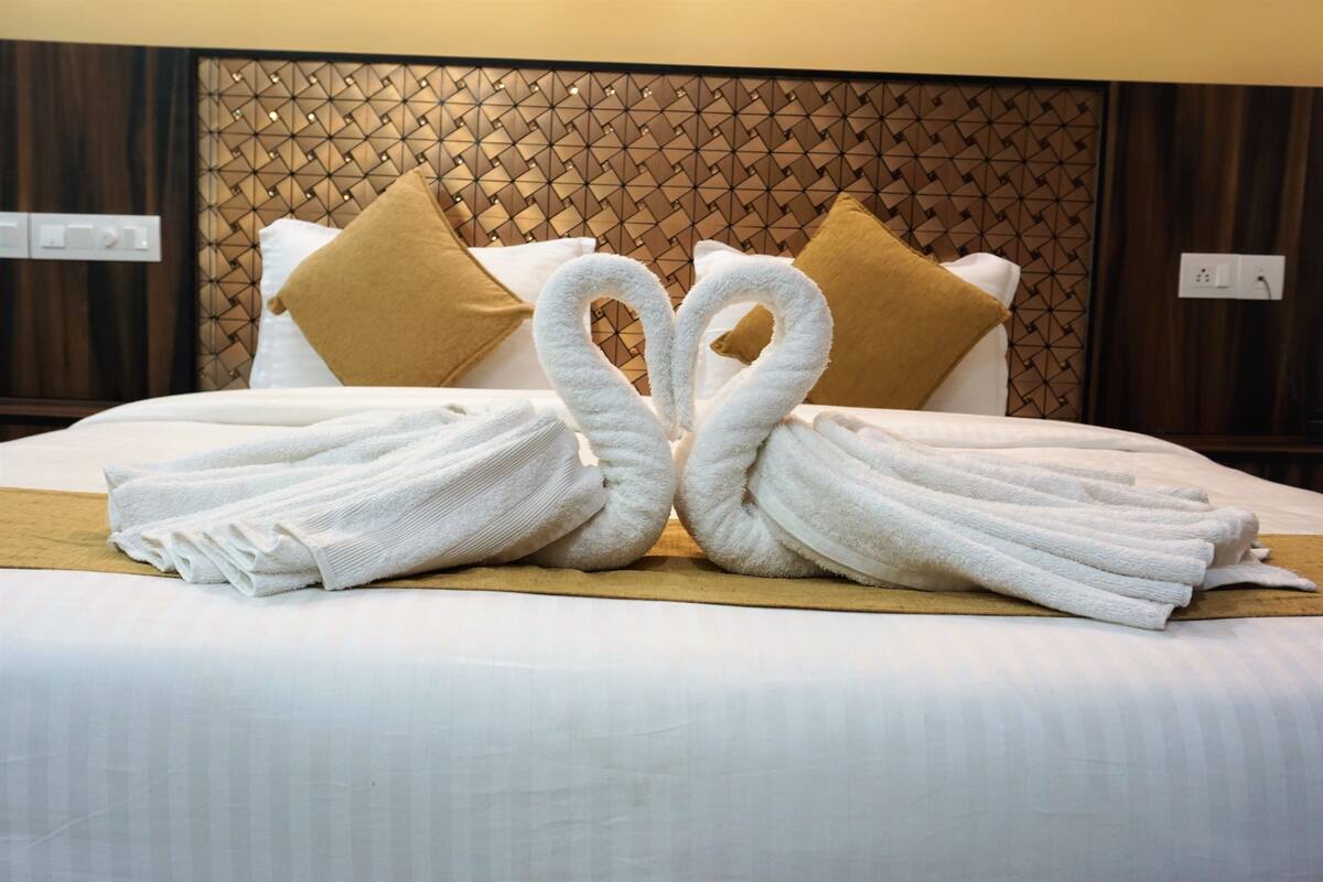 A bed with a geometric headboard, white bedding, and two towels arranged in a heart shape.