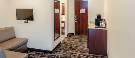 Un vestíbulo de hotel con un sofá, un televisor de pantalla plana montado en la pared, una alfombra estampada y un mostrador de recepción con una cafetera.