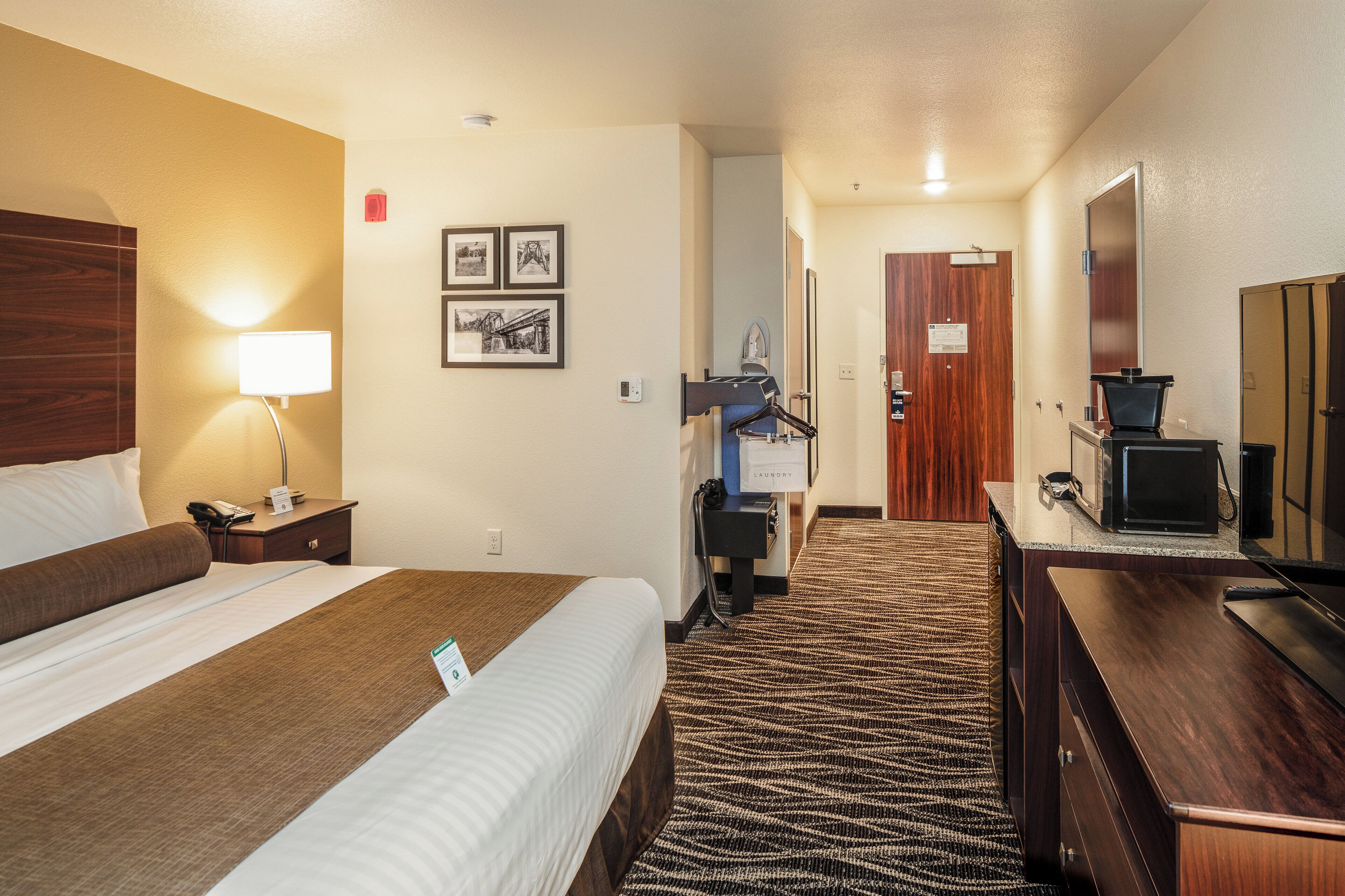 Habitación de hotel con una cama, un escritorio con computadora, un espejo y una puerta que da a otra habitación.