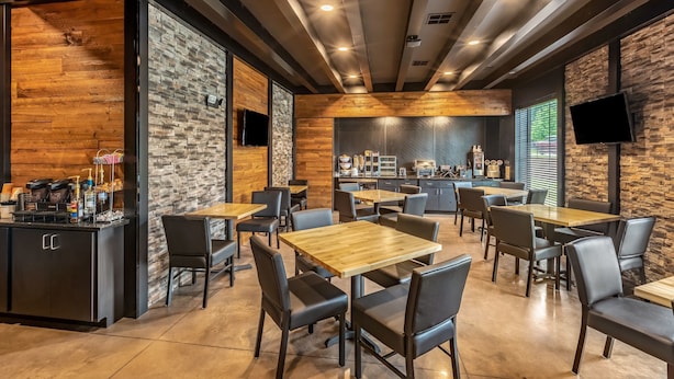 A modern cafe with wooden tables and black chairs, a kitchen area in the background, and a TV mounted on the wall. - Cobblestone Inn & Suites - Fairfield Bay (Fairfield Bay)