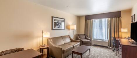 Habitación de hotel con sofá, sillón, escritorio y un ventanal con cortinas.