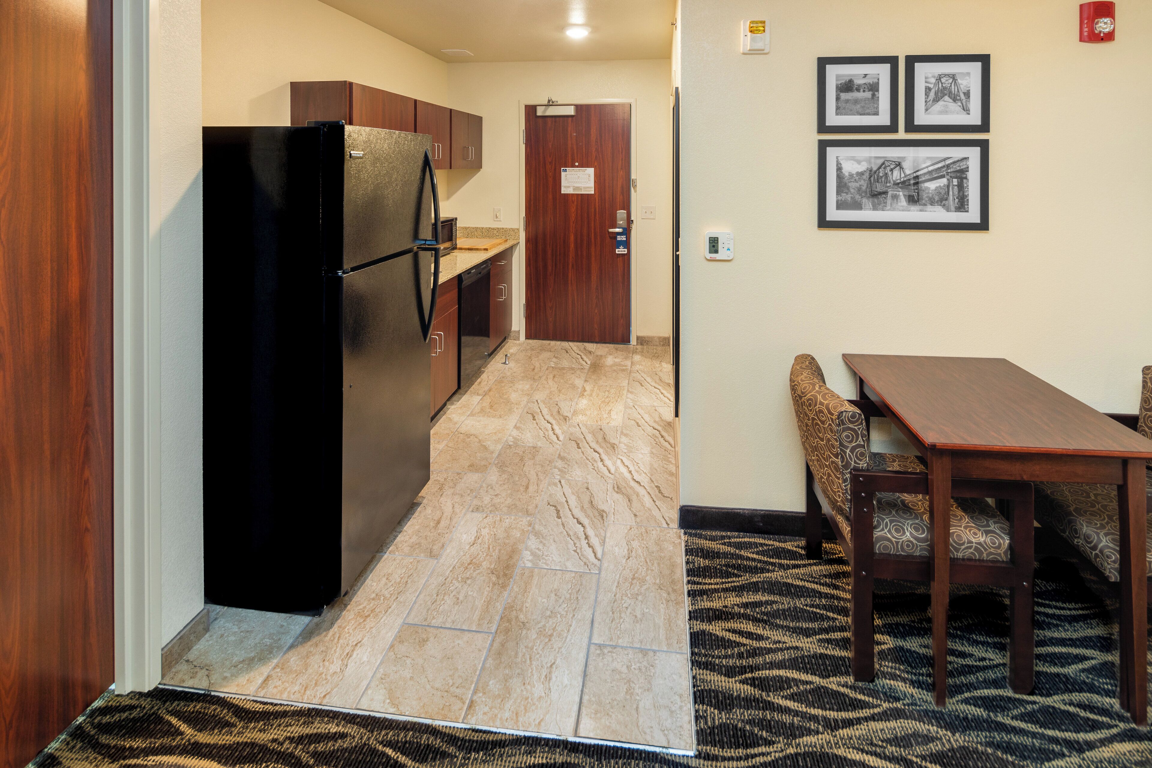 A compact kitchen with a refrigerator, wooden cabinets, and a marble-tiled floor. There is a dining area with a table and patterned bench.