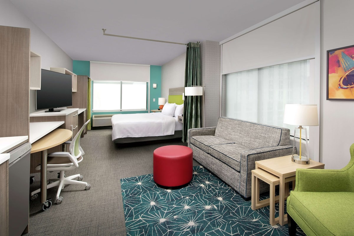 A hotel room with a bed, a desk with a computer, a red ottoman, a green armchair, and a patterned rug.