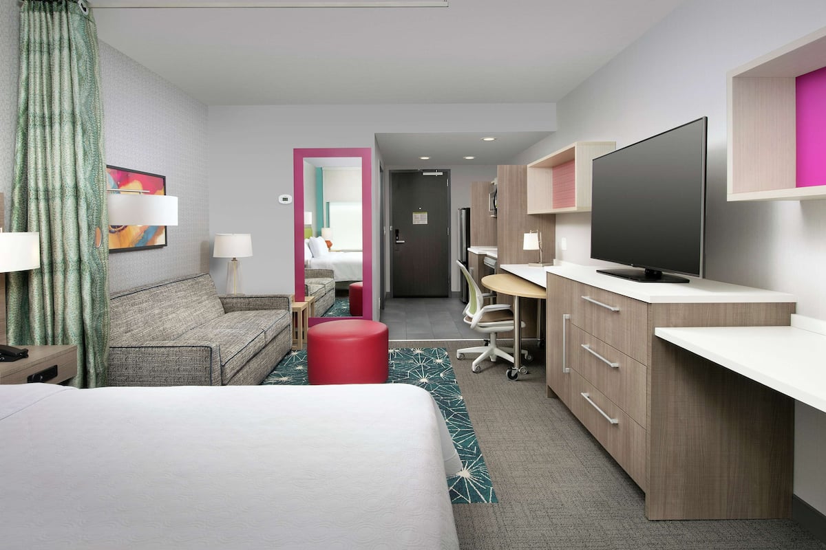 A hotel room with a bed, a desk, a TV, a chair, and a red ottoman.