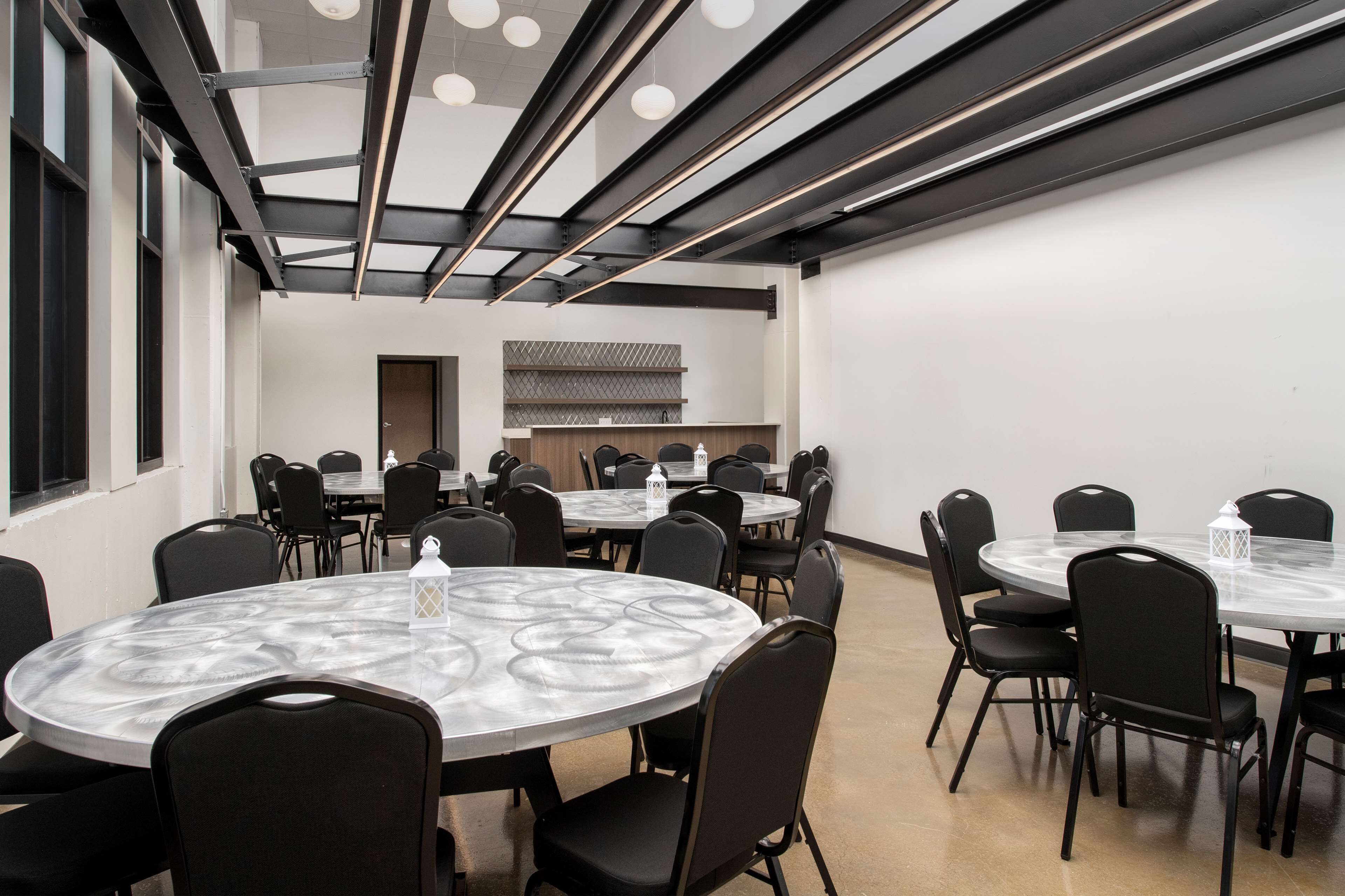 A conference room with round tables and black chairs, a kitchen area in the background, and a ceiling with exposed beams and lighting fixtures.
