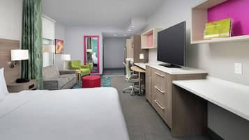 A modern hotel room with a bed, a desk, a TV, and a seating area with chairs and a red ottoman.