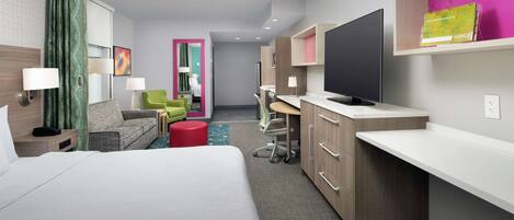 A modern hotel room with a bed, a desk, a TV, and a seating area with chairs and a red ottoman.
