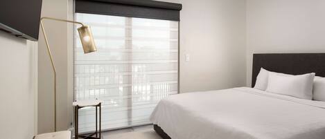 A modern hotel room with a large bed, a bedside table, a floor lamp, and a window with blinds.