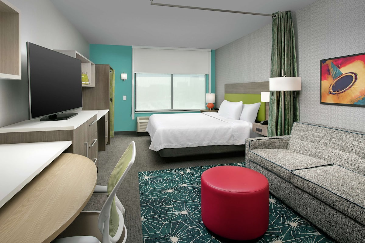 A hotel room with a bed, a sofa, a red ottoman, a TV, and a desk.