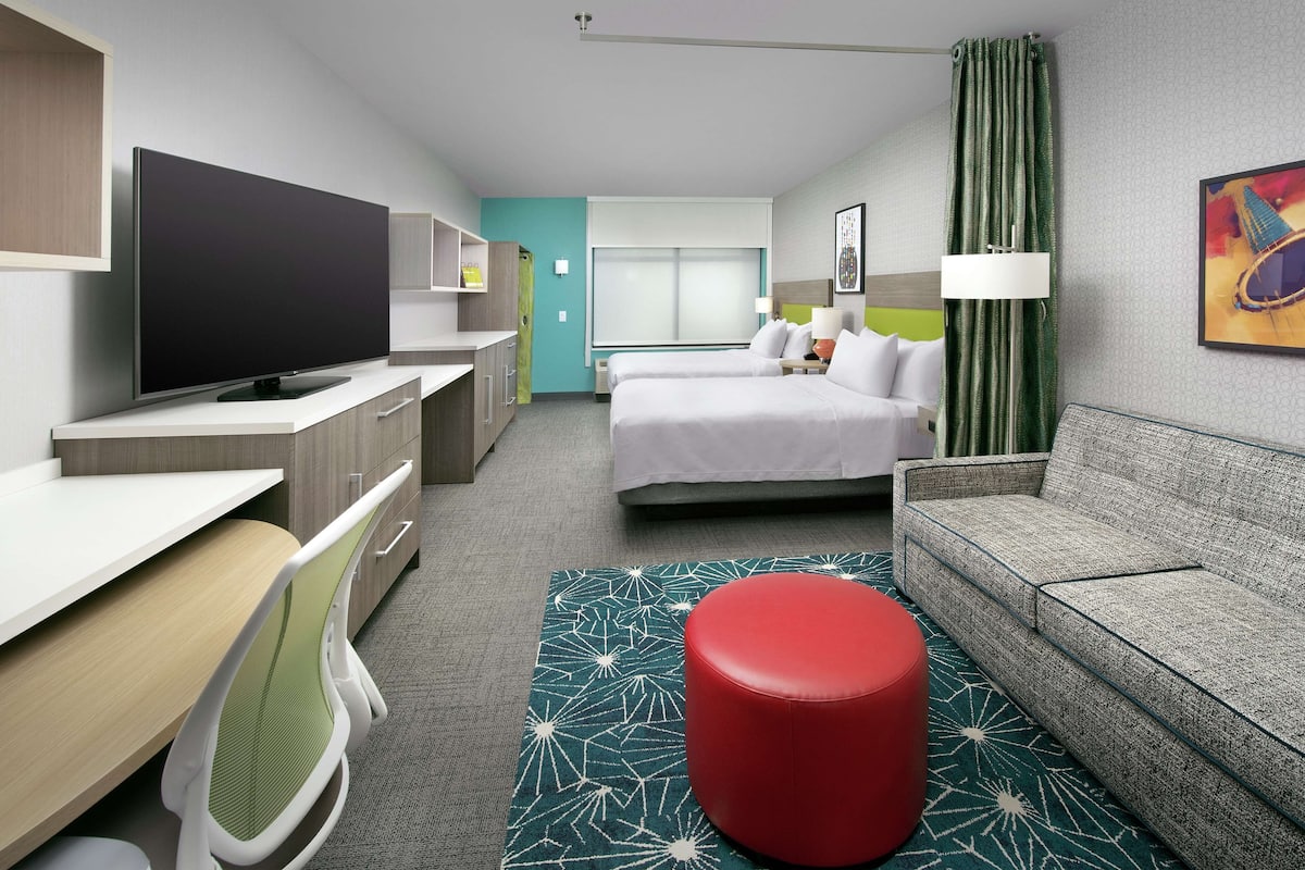 A hotel room with a bed, a sofa, a TV, a desk, and a red ottoman.