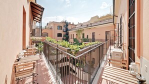 A narrow balcony with wooden furniture and potted plants, overlooking a residential area with buildings and a clear sky. - Peroni Apartment (Rome)