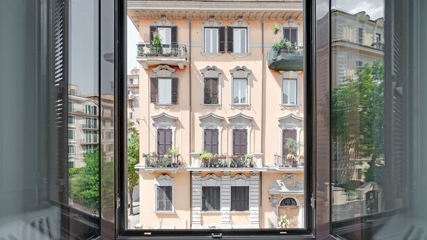 A view through a window of a building with multiple balconies, ornate architectural details, and a clear sky. - Peroni Apartment (Rome)
