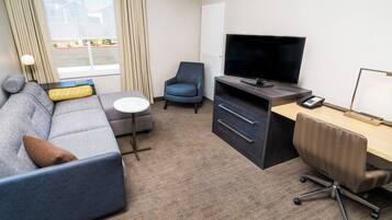 A hotel room with a gray sofa, a small round table, a blue armchair, a TV on a wooden stand, a desk with a chair, and a window with curtains.