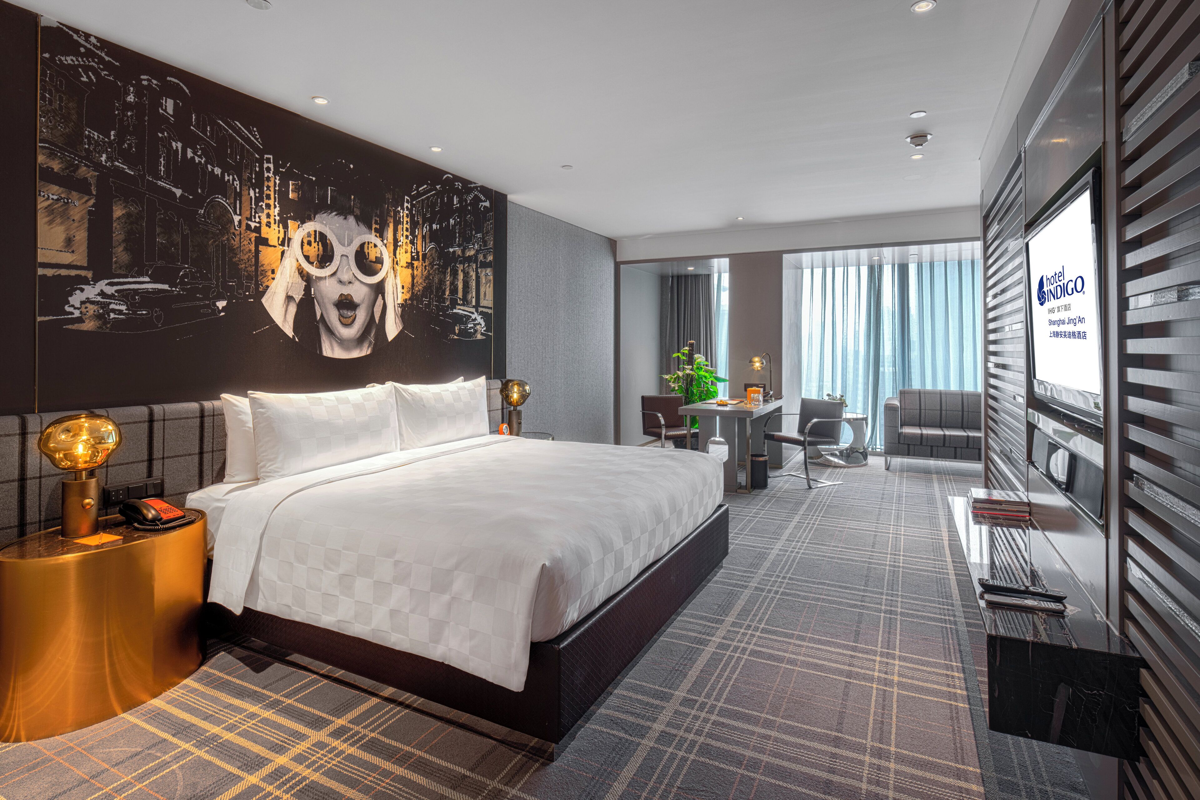 A hotel room with a large bed, a desk, a chair, a small table, and a large window.