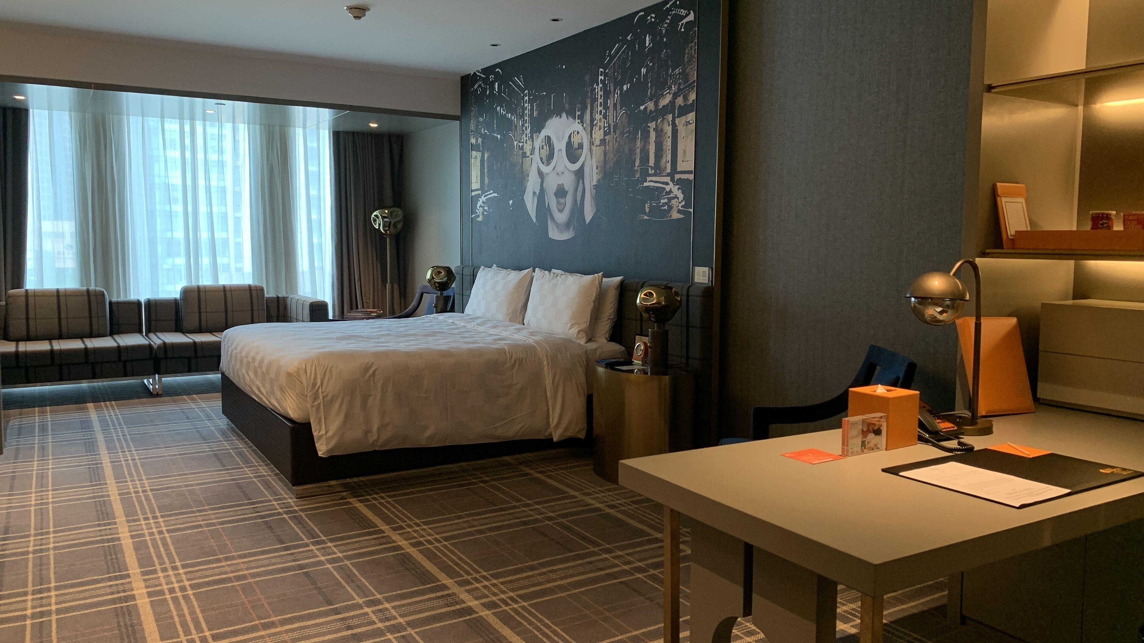 A hotel room with a large bed, a desk with a lamp, a sofa, and a wall with a cityscape mural.