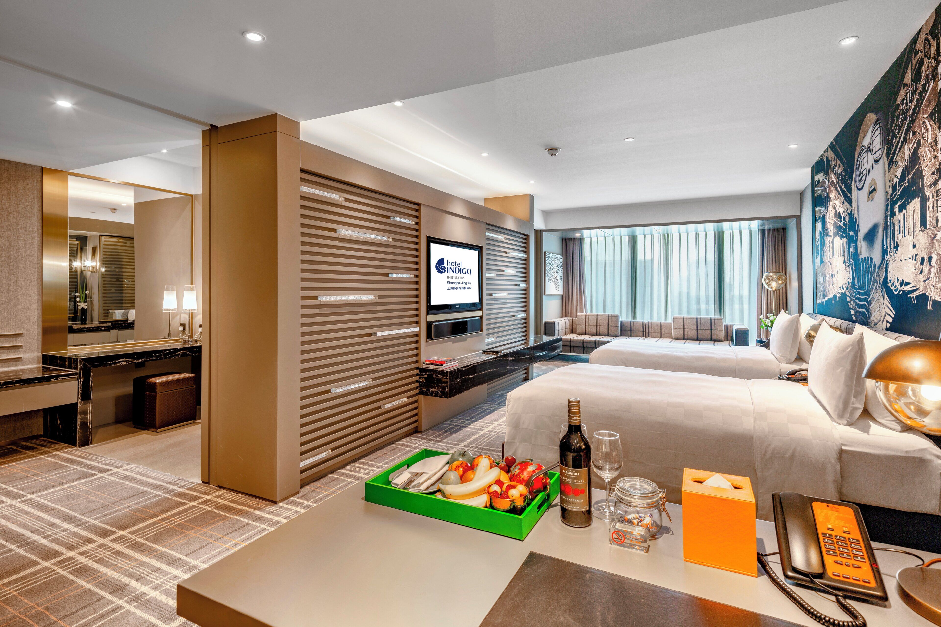A hotel room with a bed, a desk, a television, and a dining area with a tray of fruit.