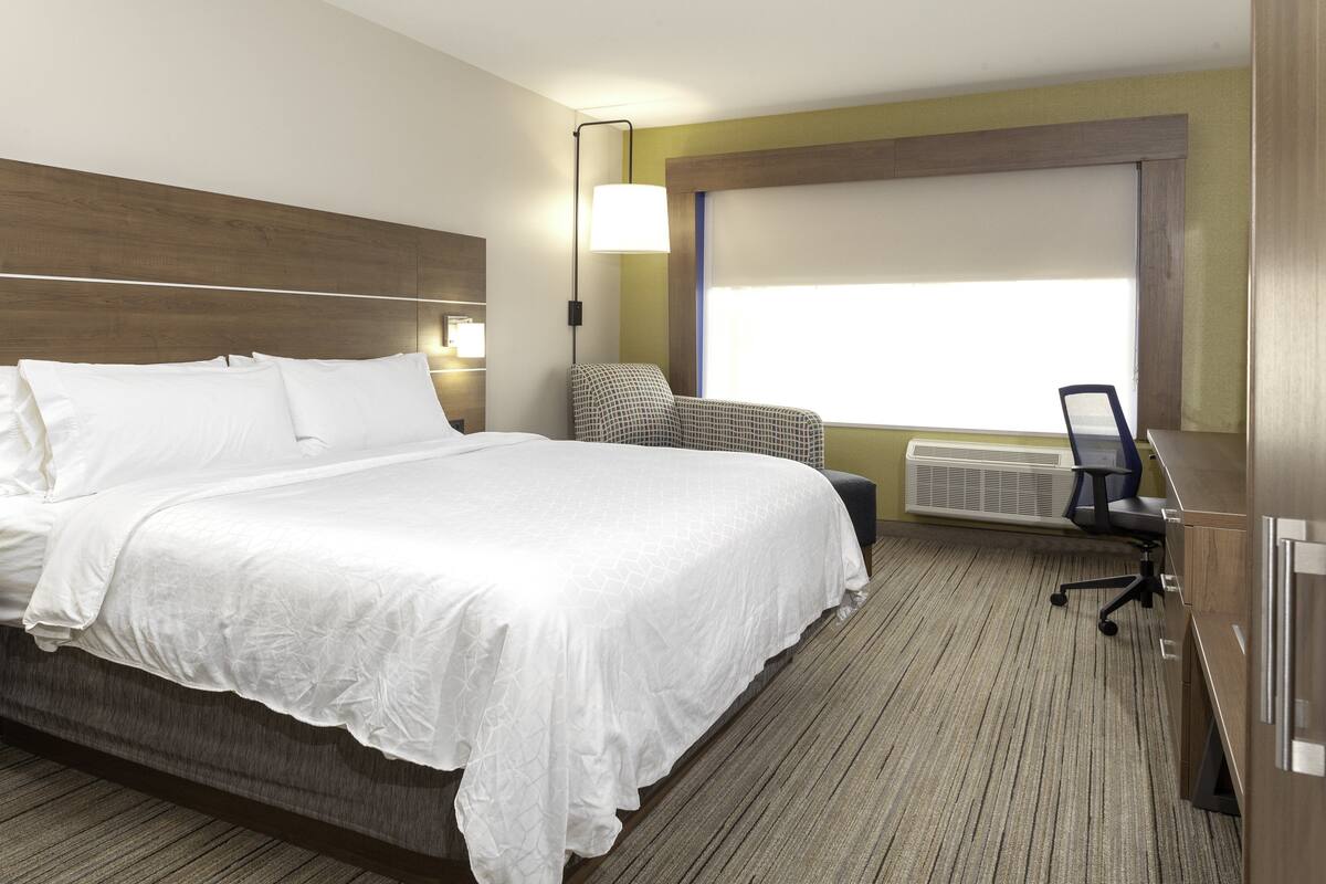 Habitación de hotel con una cama grande, un escritorio con silla, una lámpara y una ventana con persianas.
