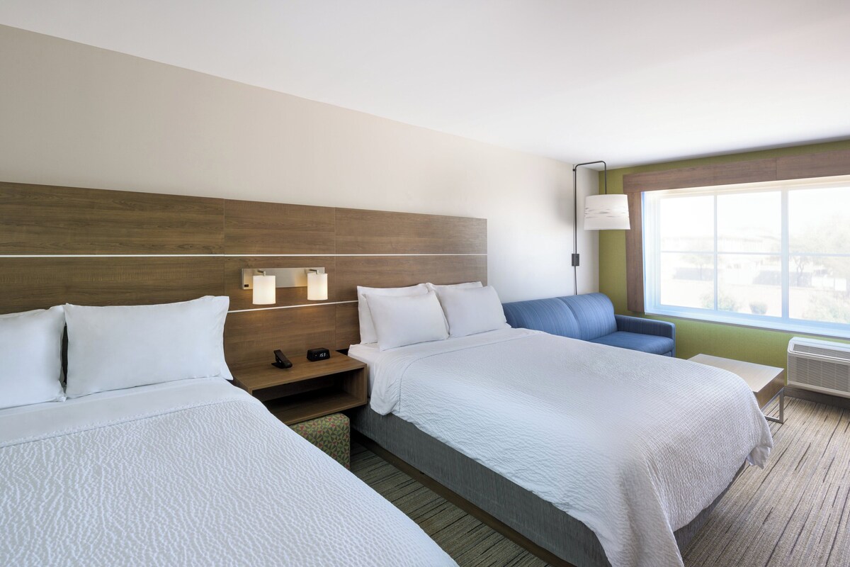 A hotel room with two beds, a nightstand, a desk, a chair, and a large window.