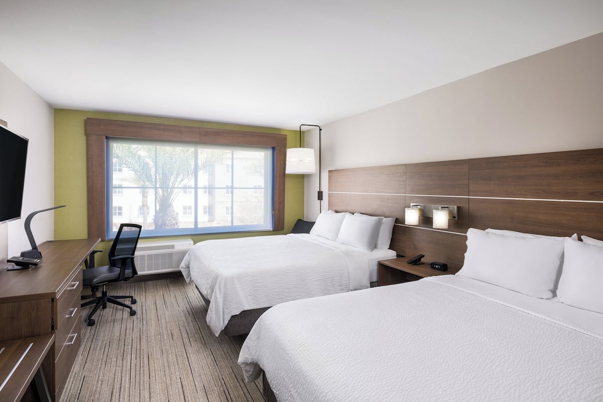 Habitación de hotel con dos camas, un escritorio, una silla, un televisor y una ventana con vista a edificios.