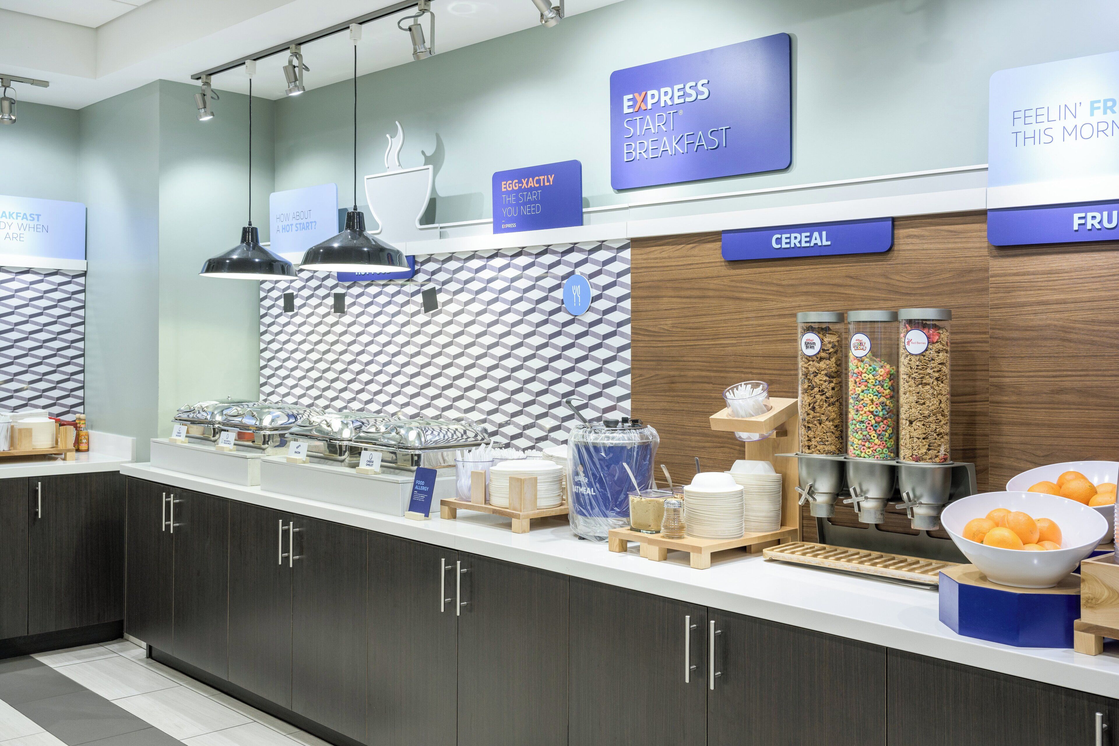 Una barra de desayuno con secciones identificadas para cereales y fruta, una máquina de café y una variedad de alimentos para el desayuno a la vista.