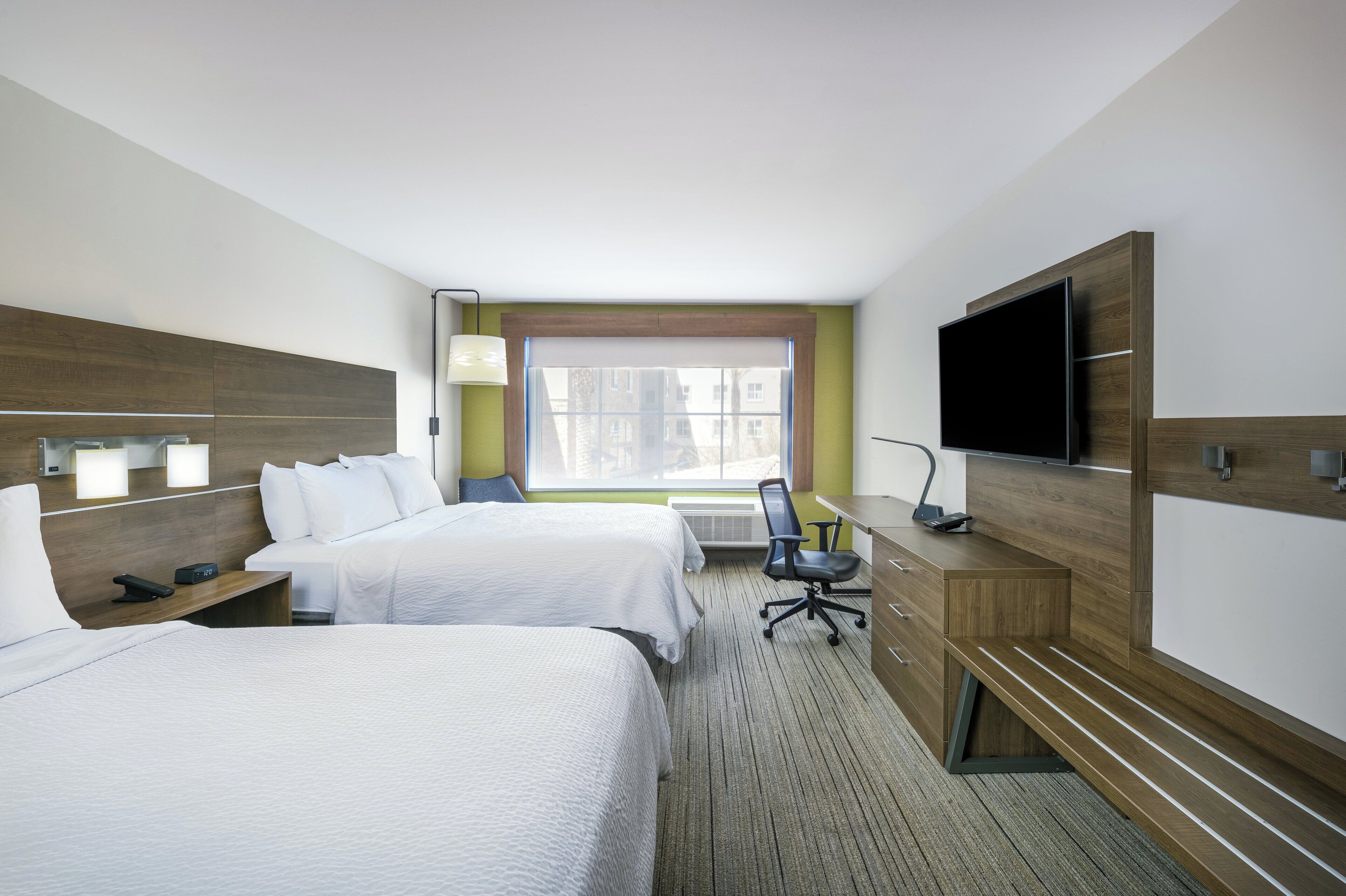 Habitación de hotel con dos camas, un escritorio, una silla, un televisor y una ventana con vista a edificios.