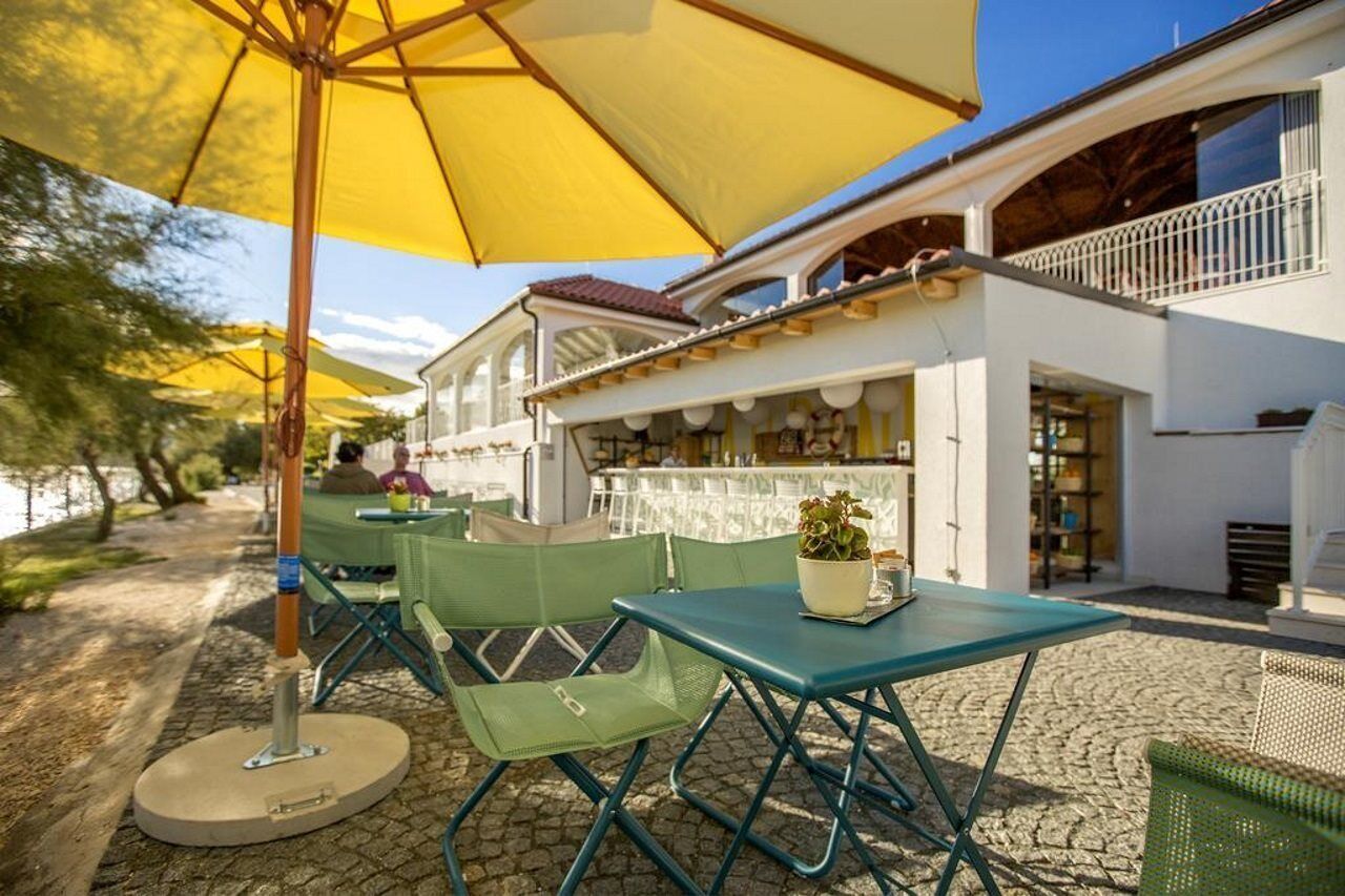An outdoor seating area with green chairs and a yellow umbrella, a white building with a balcony, and a river in the background.