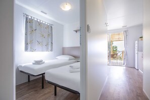 A bedroom with two beds, a window with a floral curtain, a bedside table, and a view of the outside balcony. -  Amadria Park Camping Trogir - Apartments (Seget)