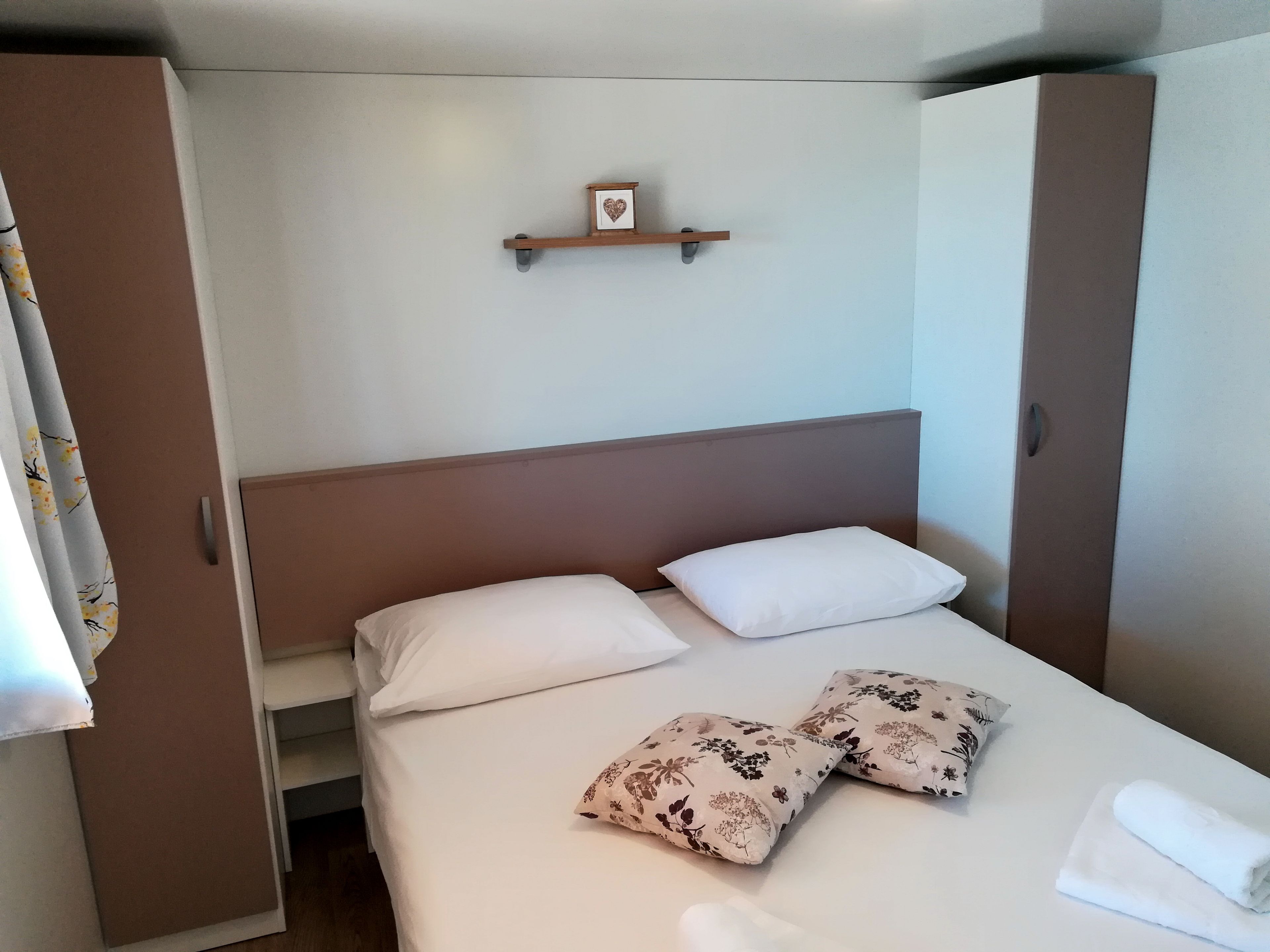 A hotel room with a bed, pillows, a bedside table, and a small shelf with a heart-shaped decoration.