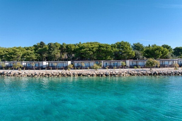 A waterfront hotel with multiple rooms, a rocky shoreline, and dense greenery in the background. -  Amadria Park Camping Trogir - Apartments (Seget)