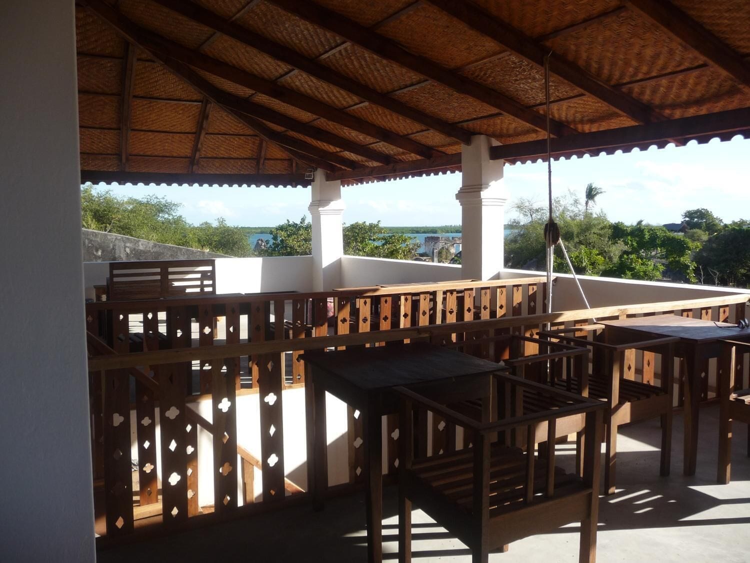 An outdoor patio with wooden tables and chairs, a thatched roof, and a view of the sea.
