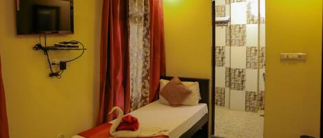 A small, single-bed room with a TV mounted on the wall, a bed with a red blanket and pillows, a curtain, and a door leading to a bathroom.