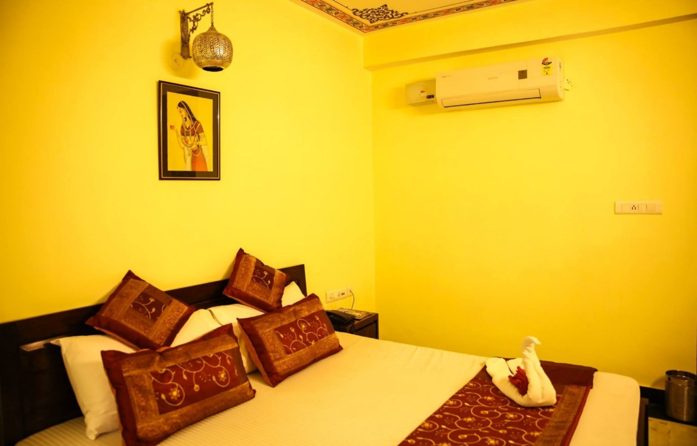 A hotel room with a bright yellow wall, a bed with brown and white pillows, a framed picture on the wall, and an air conditioning unit.