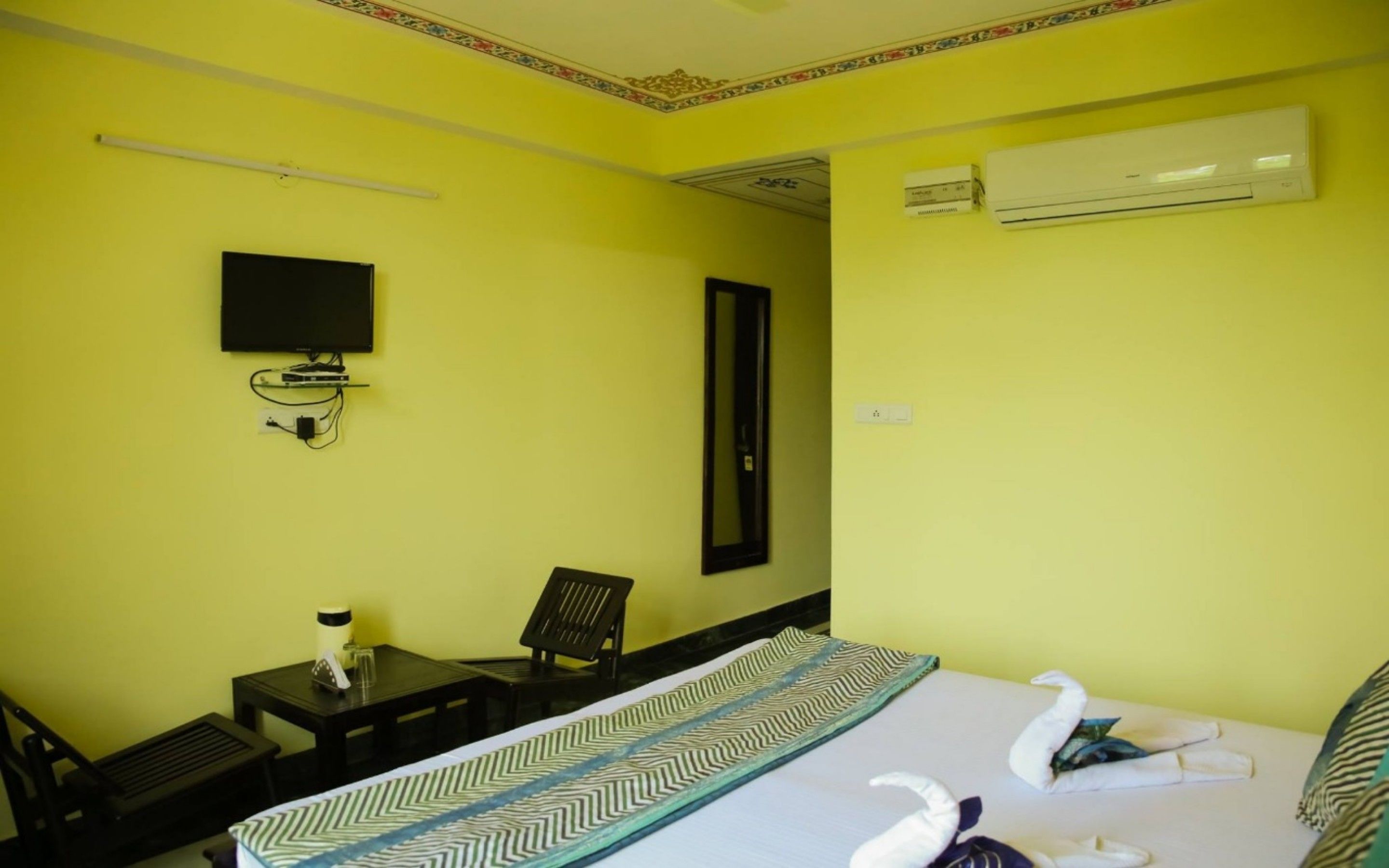 A hotel room with a bed, a TV mounted on the wall, an air conditioning unit, and a desk with a chair.